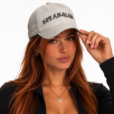 A woman with long red hair wearing a light gray baseball cap with 'OUTANGLED' embroidered in black gothic lettering, posed against a solid white background.
